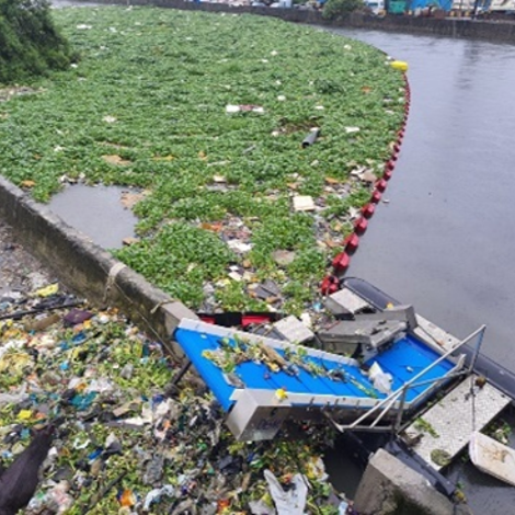 Waterway clogged with trash and plants, featuring a DESMI aquatic waste collection system.