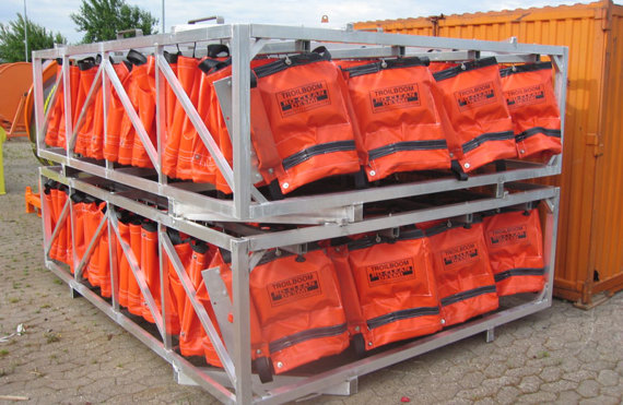 Orange TROILBOOM oil spill barriers stacked on metal frames outdoors.