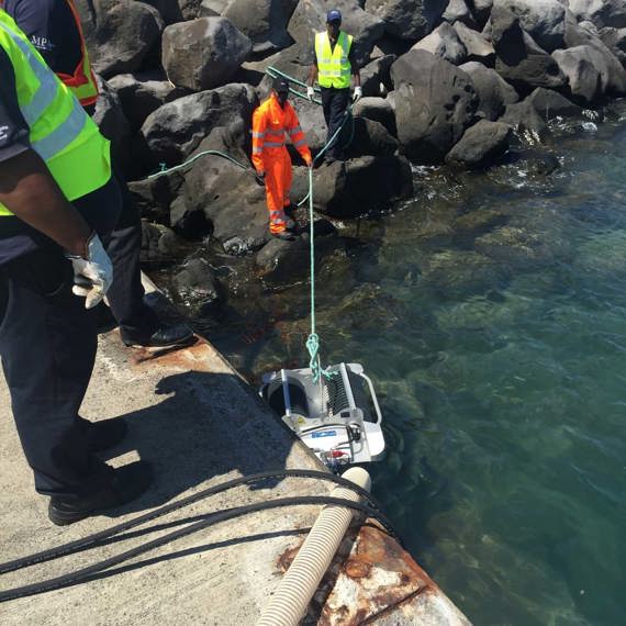 Workers deploying DESMI equipment for fluid handling near rocky shoreline.