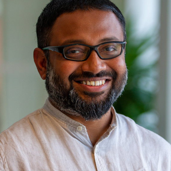 Smiling man with glasses and beard wearing a light-colored shirt indoors.