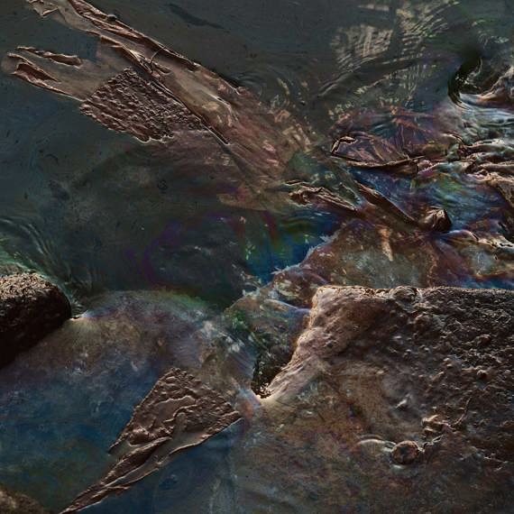 Oil slick with iridescent colors on water, surrounded by rocks and debris.