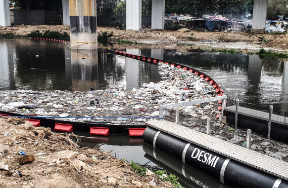 DESMI boom system collecting debris and waste from a polluted river under a bridge.