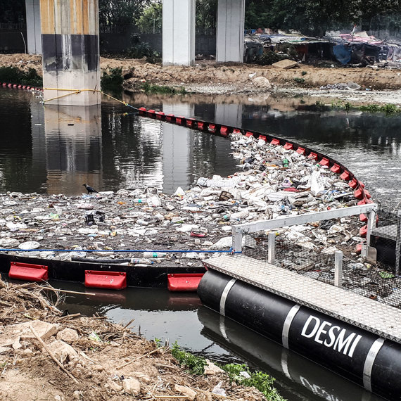 DESMI boom system collecting debris and waste from a polluted river under a bridge.