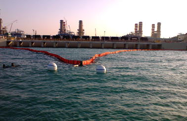 Industrial facility with floating barrier on water, two divers visible in foreground.