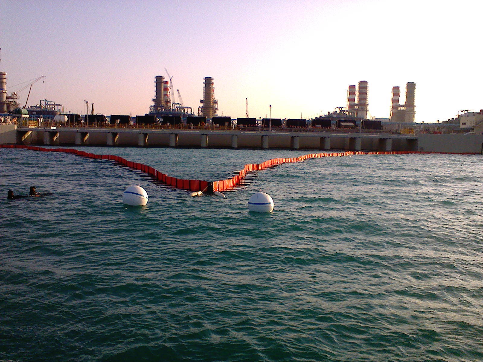 Industrial facility with floating barrier on water, two divers visible in foreground.