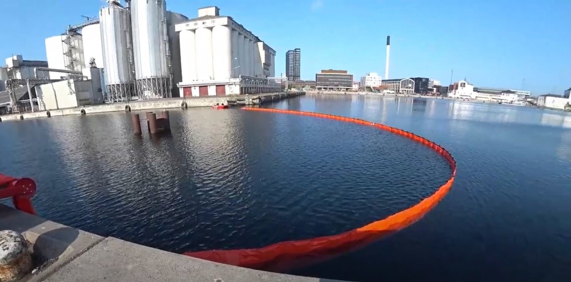 Orange oil containment boom in an industrial harbor setting under a clear sky.