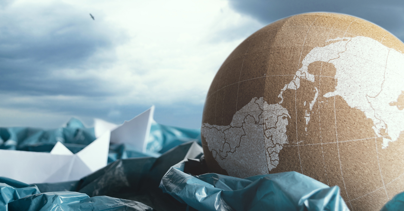 Globe amidst plastic waste and paper boats under a cloudy sky.