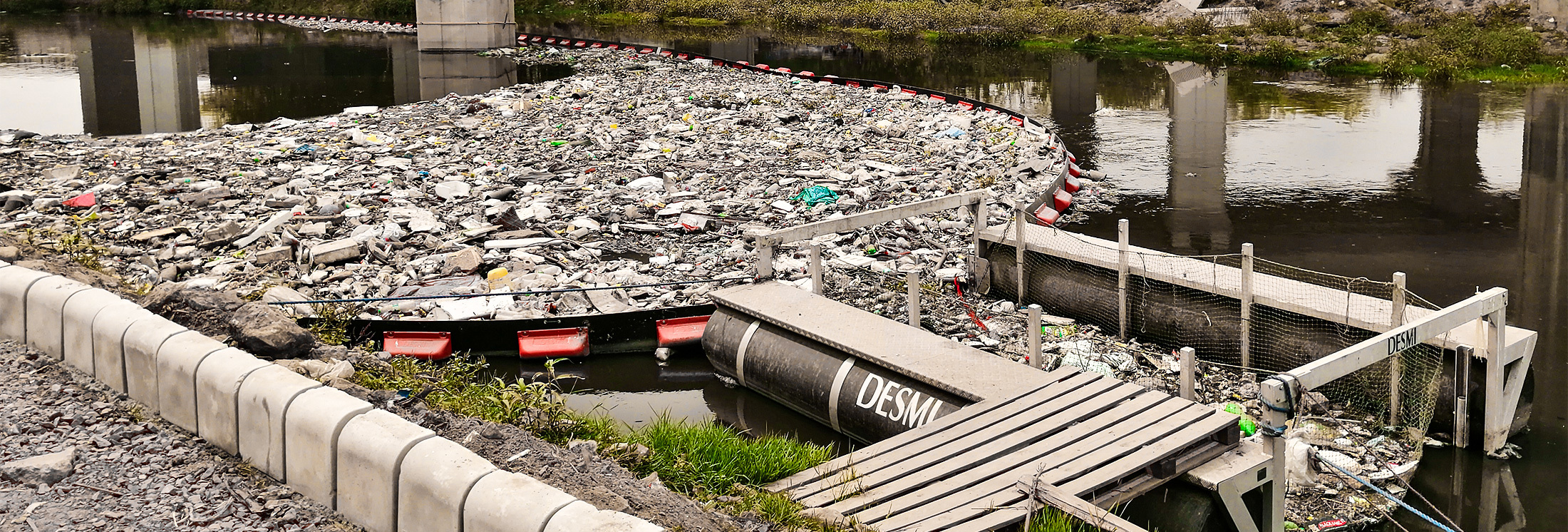 DESMI system collecting floating debris in a polluted waterway under a bridge.