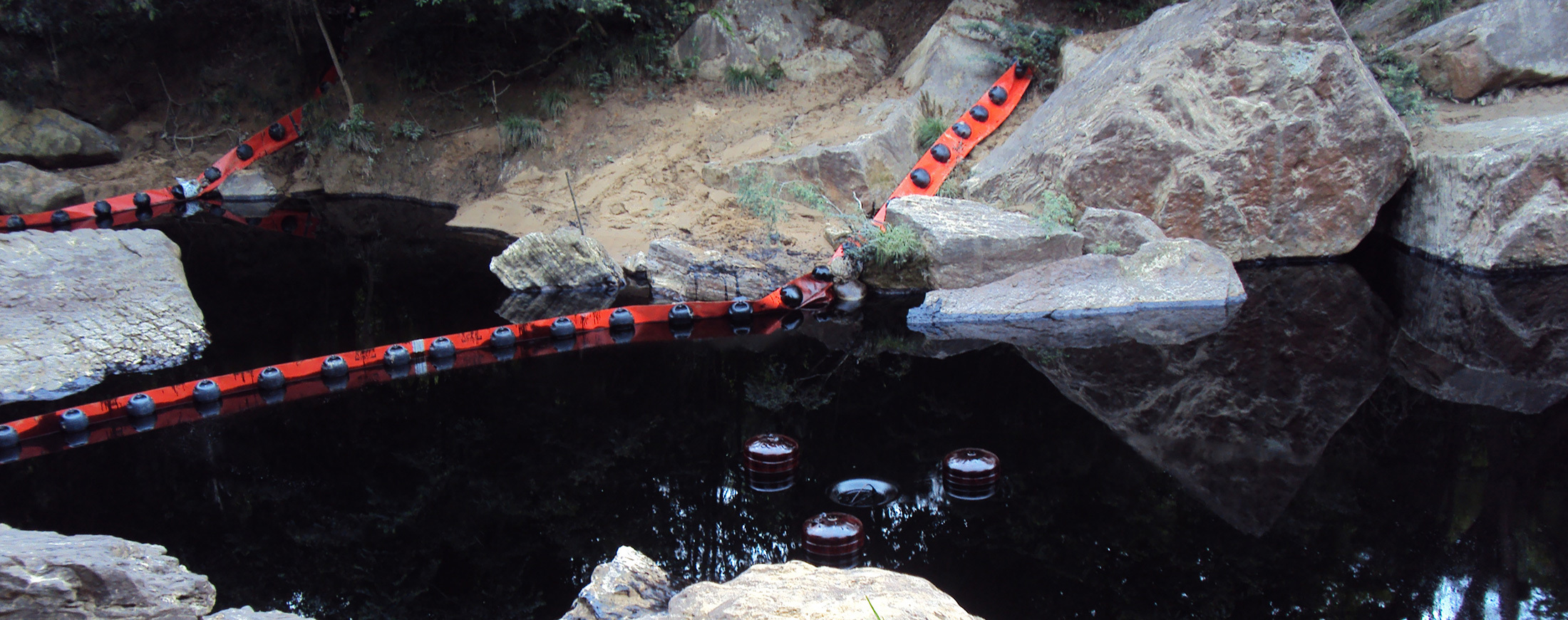 Red oil containment boom in rocky, forested waterway for spill control.