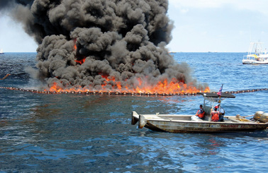 Boat near ocean fire containment with thick black smoke rising.