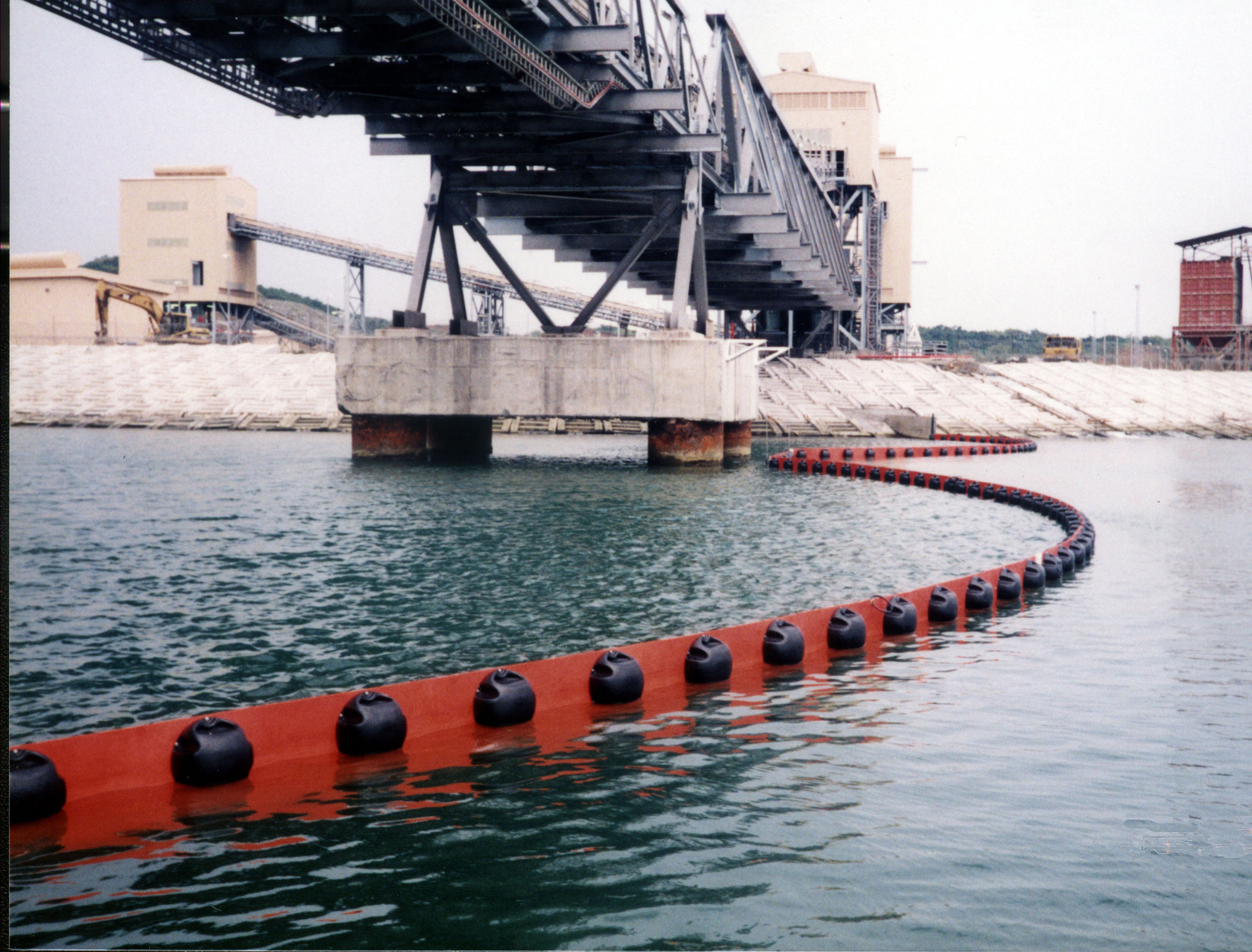 Red floating barrier in water near industrial facility and bridge.