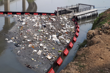 DESMI barrier containing floating debris on a polluted river surface.