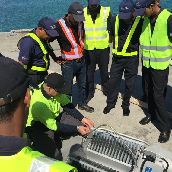 Group of workers in safety vests observing equipment demonstration near the water.