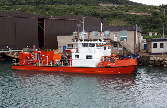 Orange and white vessel "Fagio II" docked near industrial buildings by green hillside.