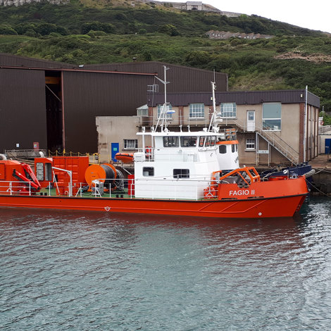 Orange and white vessel "Fagio II" docked near industrial buildings by green hillside.