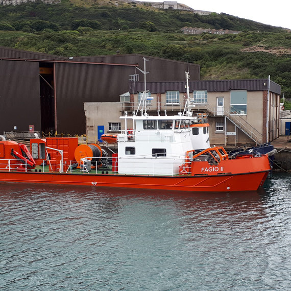 Orange and white vessel "Fagio II" docked near industrial buildings by green hillside.